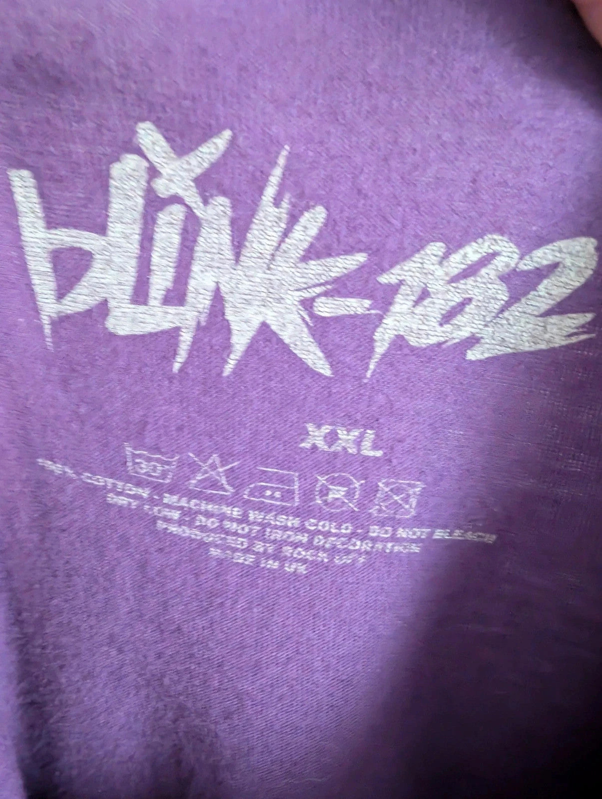 Blink-182 purple graphic tee featuring smiley face logo with neon pink outlining and drip detailing.