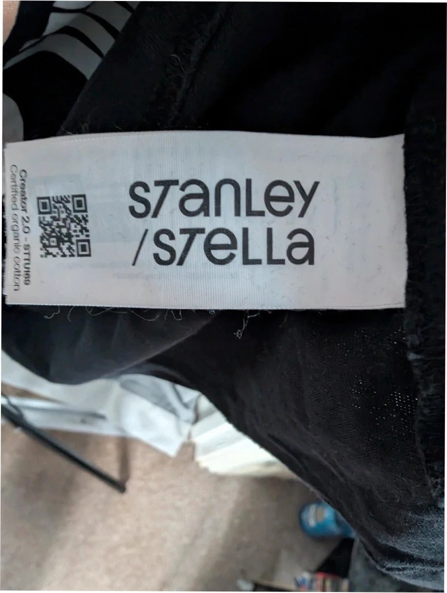 Stanley/Stella black graphic tee featuring Oasis logo and "Roll With It" beach artwork.