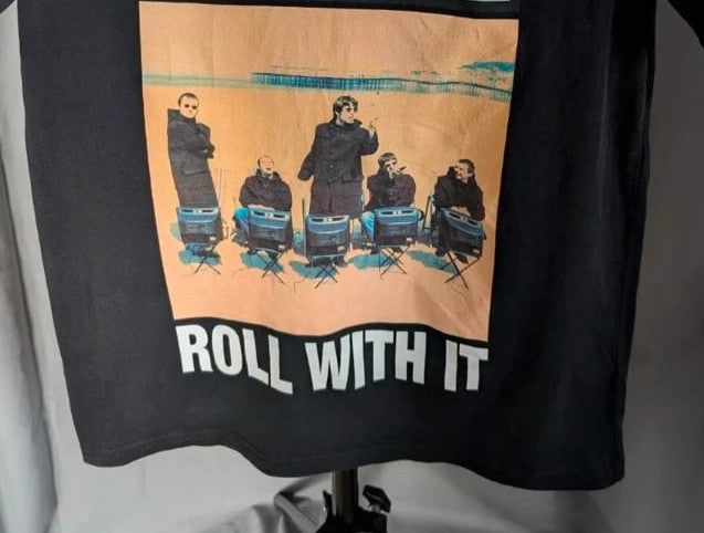 Stanley/Stella black graphic tee featuring Oasis logo and "Roll With It" beach artwork.