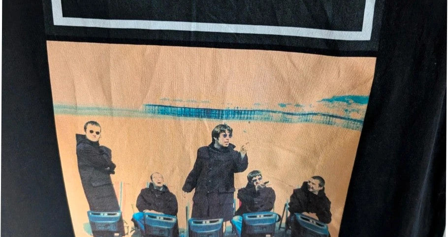 Stanley/Stella black graphic tee featuring Oasis logo and "Roll With It" beach artwork.