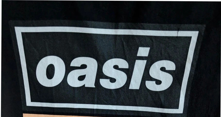 Stanley/Stella black graphic tee featuring Oasis logo and "Roll With It" beach artwork.